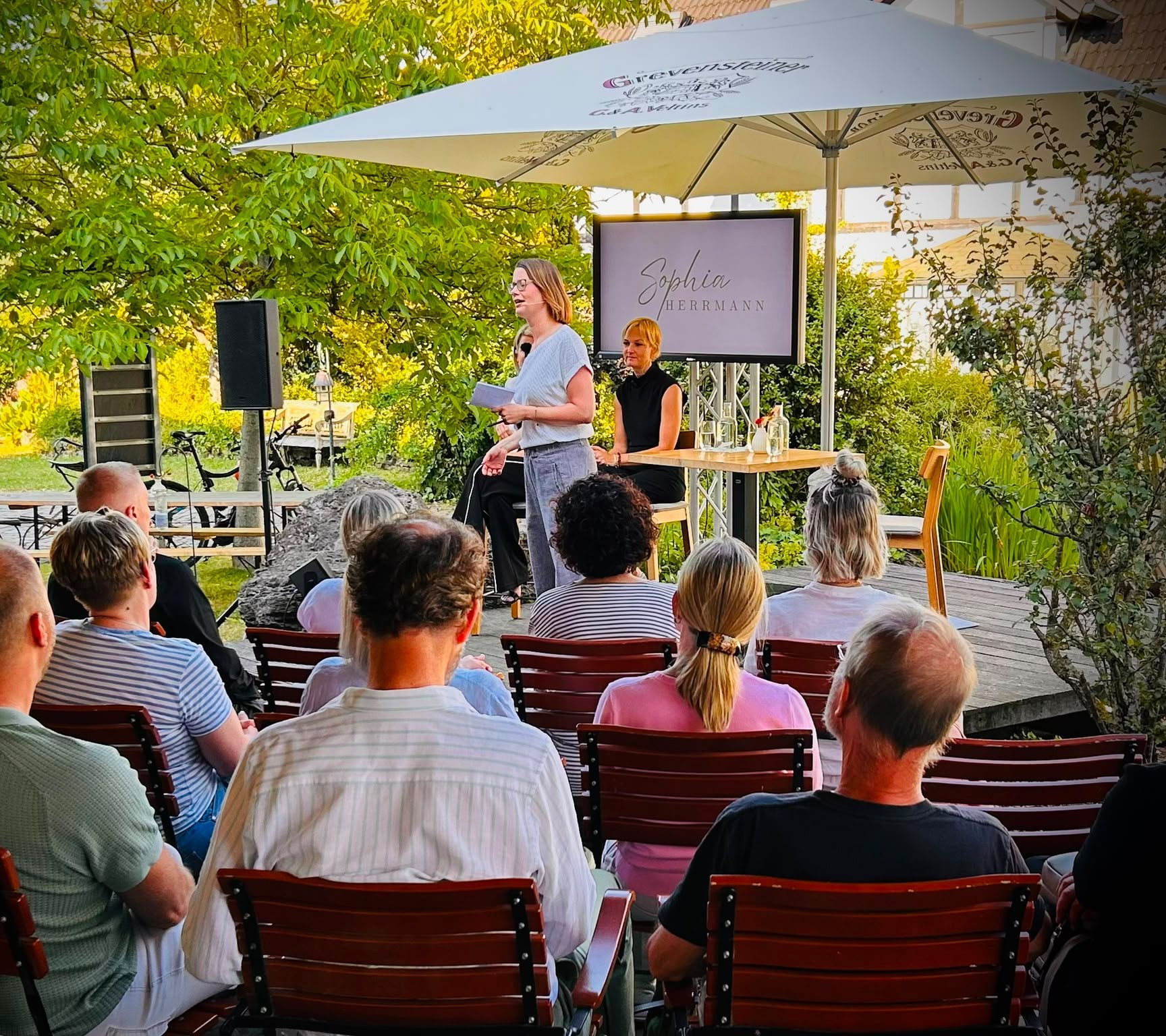 Sophia Herrmann spricht beim MindWeib-Netzwerktreffen vor einem Publikum auf einer Gartenterrasse; hinter ihr zeigt ein Bildschirm ihren Namen.